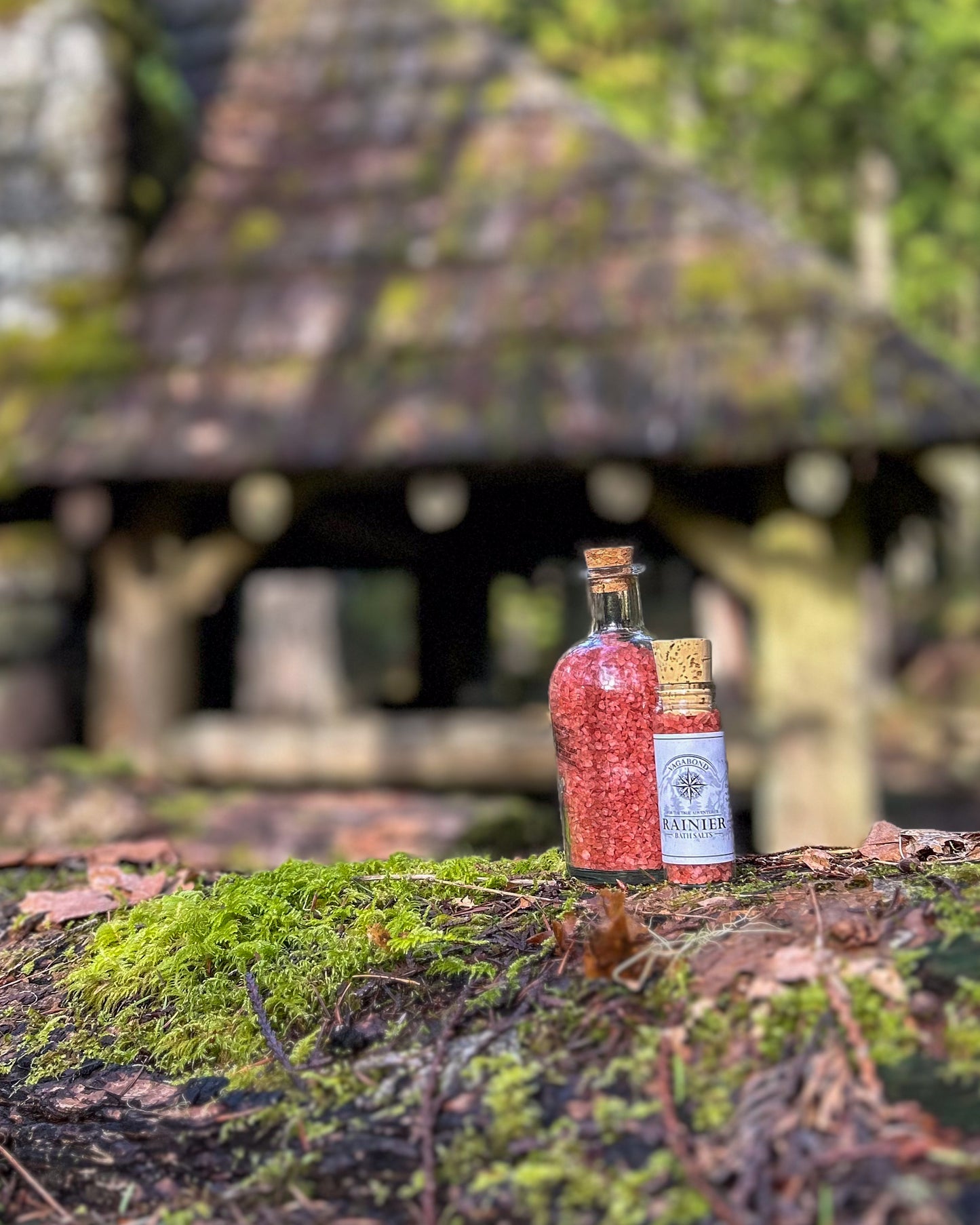 Rainier natural bath salts warm cinnamon scent in corked glass bottle displayed against a mossy cabin backdrop