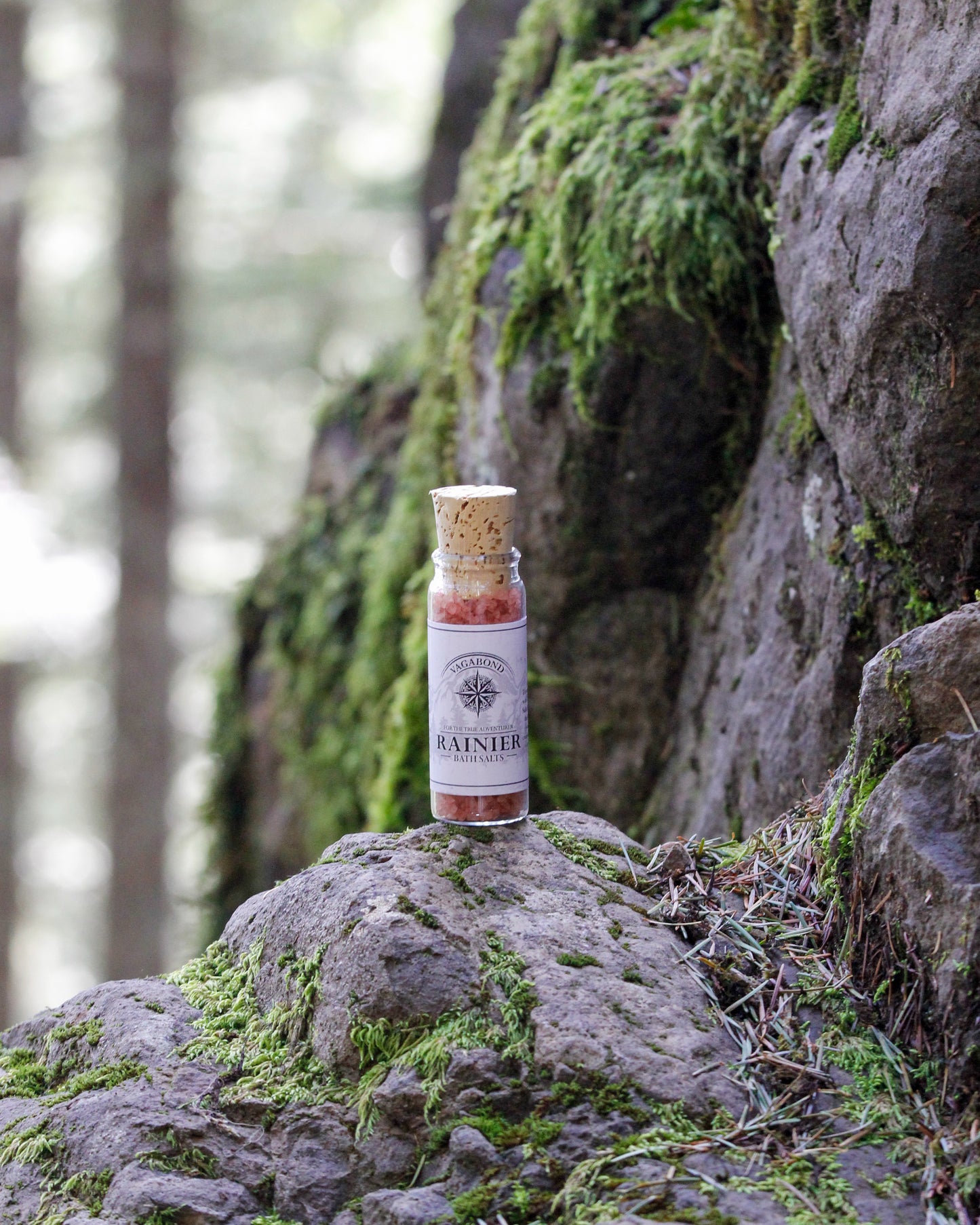 Rainier natural bath salts warm cinnamon scent in corked glass bottle displayed against a mossy mountainside backdrop