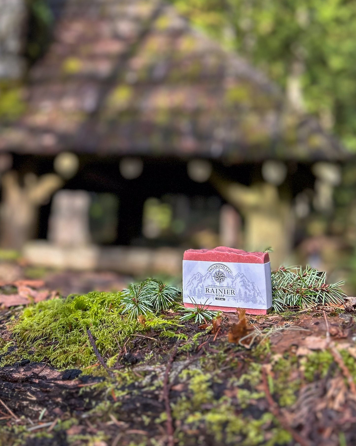 Rainier natural bar soap warm cinnamon scent on a log displayed with climbing gear against a mossy cabin backdrop.