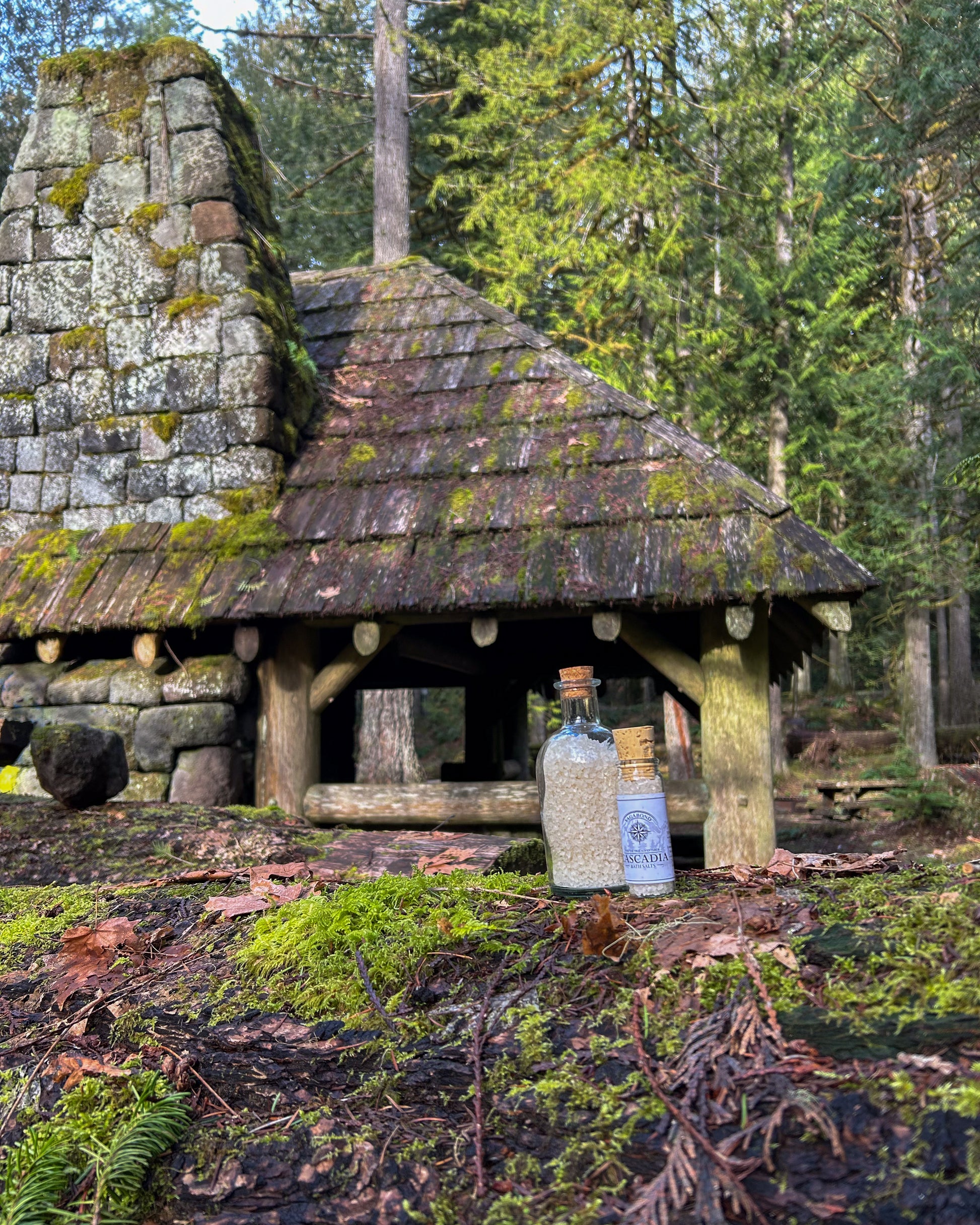 Cascadia natural bath salts lemon citrus scent in corked glass bottle displayed against a mossy cabin backdrop.