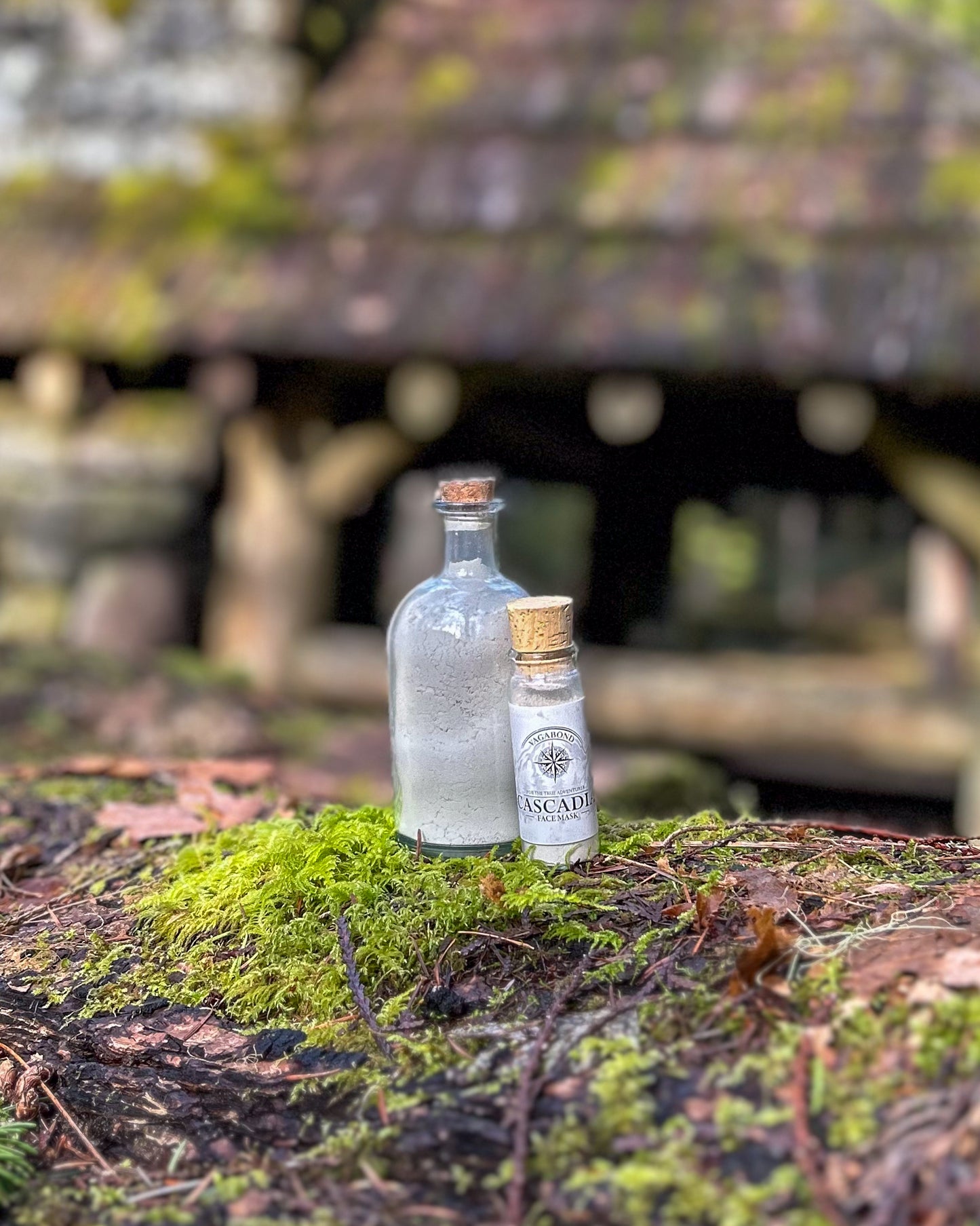 Cascadia natural clay mask in corked glass bottles displayed against a mossy cabin backdrop.