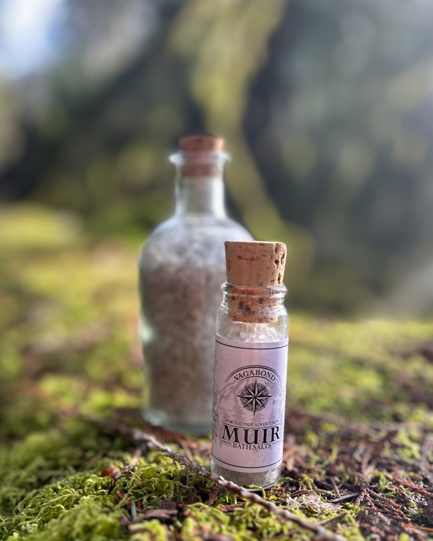 Muir natural bath salts evergreen scent in corked glass bottles displayed on a mossy log backdrop