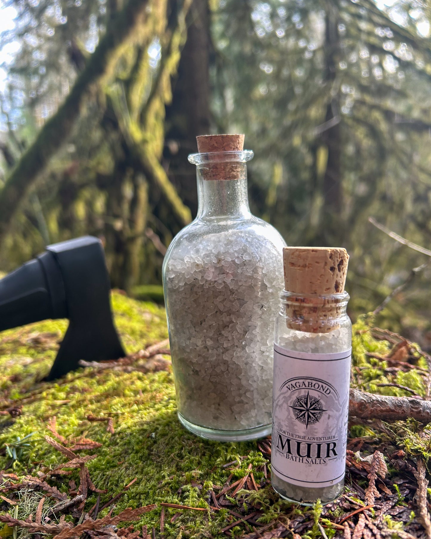 Muir natural bath salts evergreen scent in corked glass bottles displayed on a mossy log with an axe backdrop