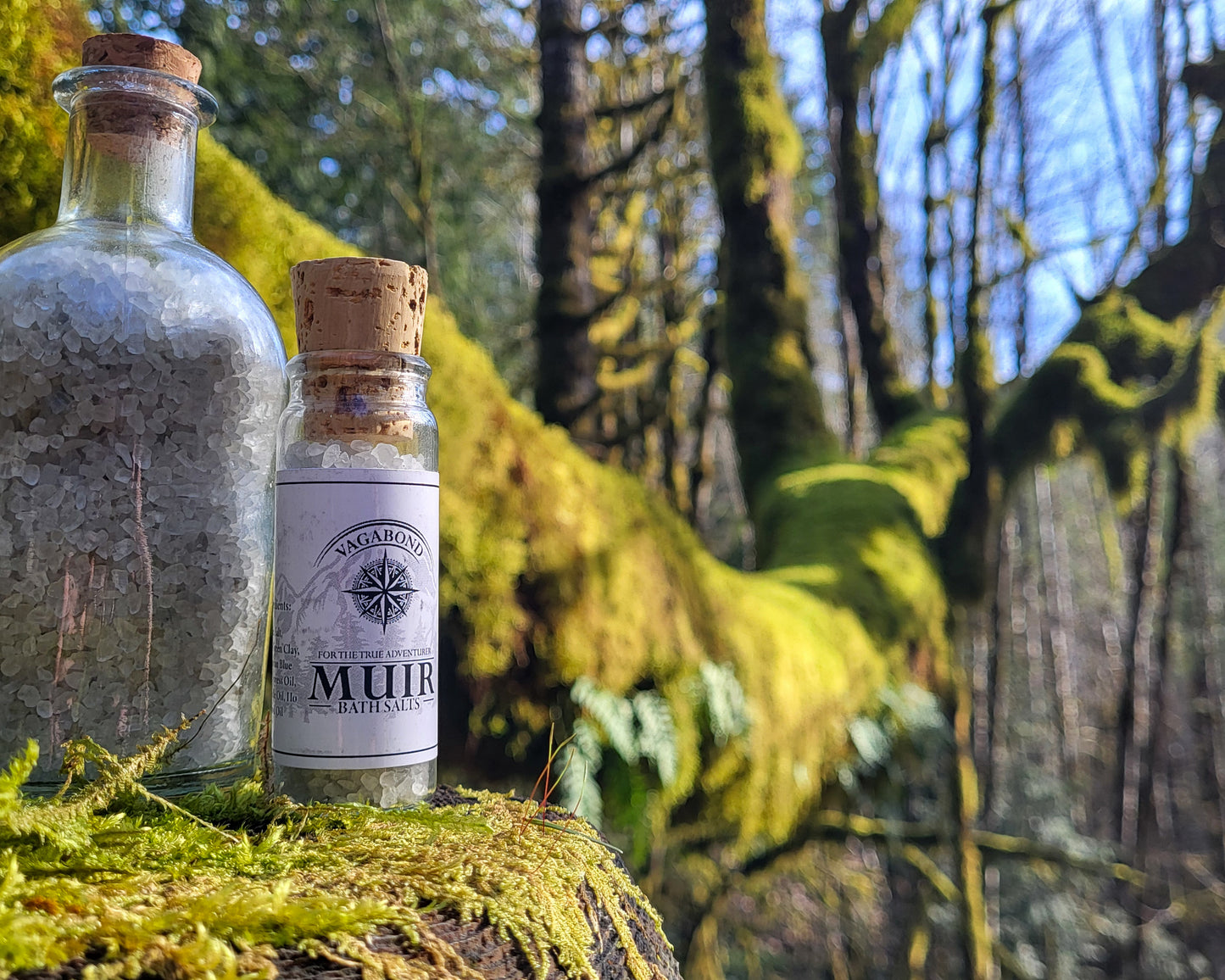 Muir natural bath salts evergreen scent in corked glass bottles displayed on a mossy branch backdrop