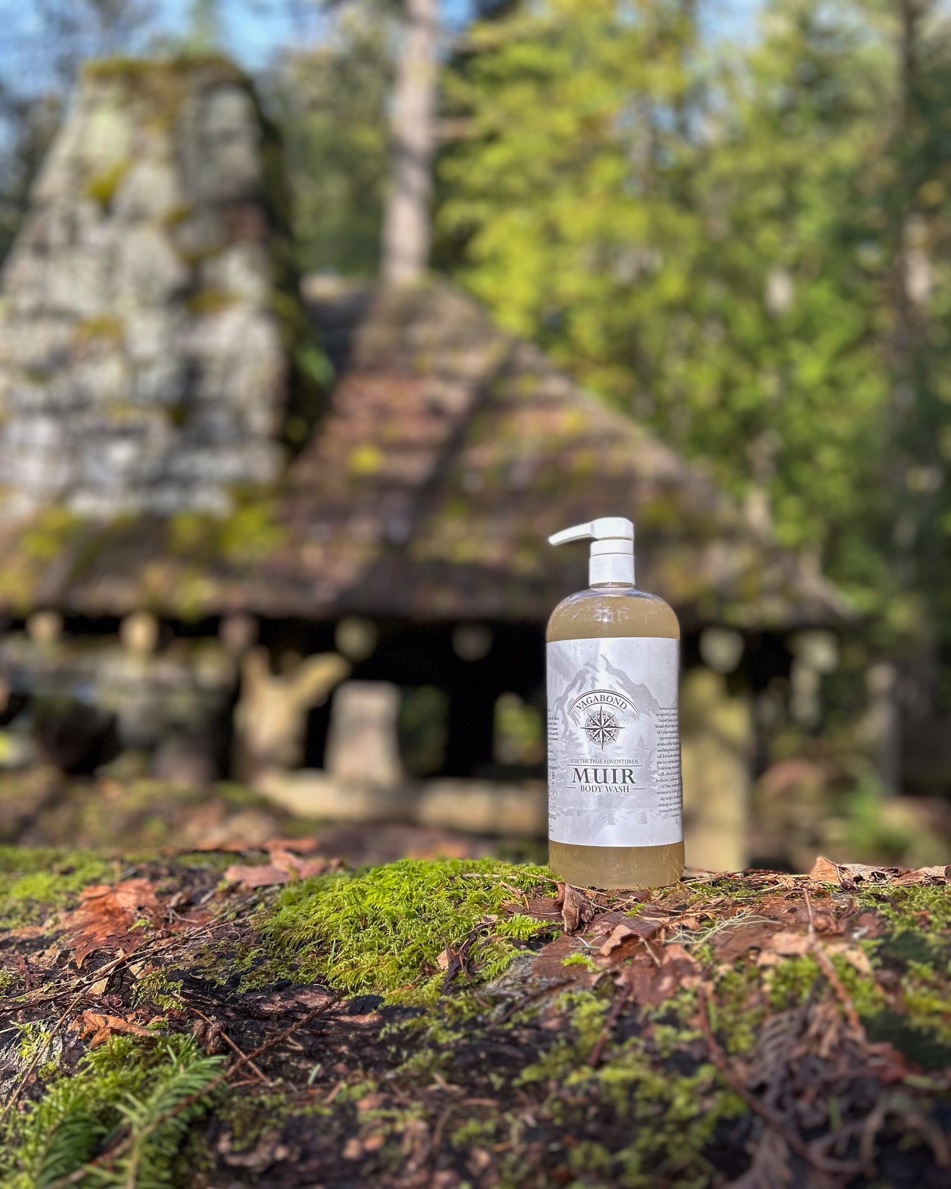 Muir natural body wash evergreen scent in corked glass bottles displayed on a mossy rock with cabin backdrop