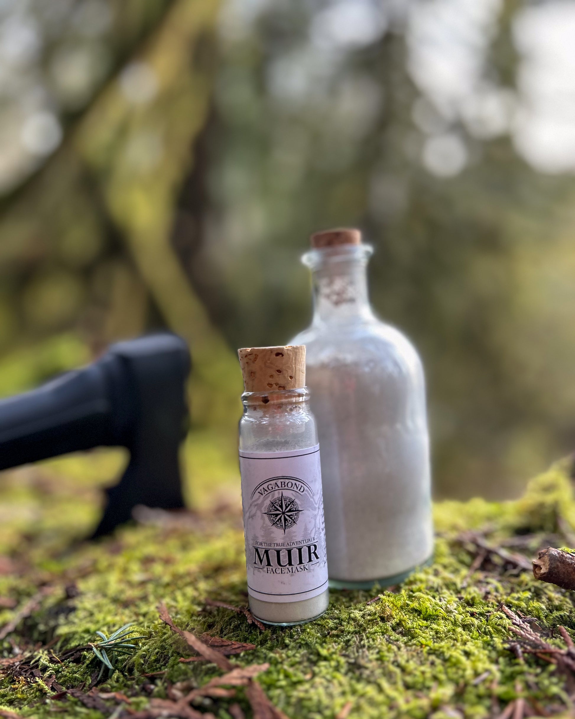 Muir natural clay mask in corked glass bottles displayed on a mossy log with axe backdrop.
