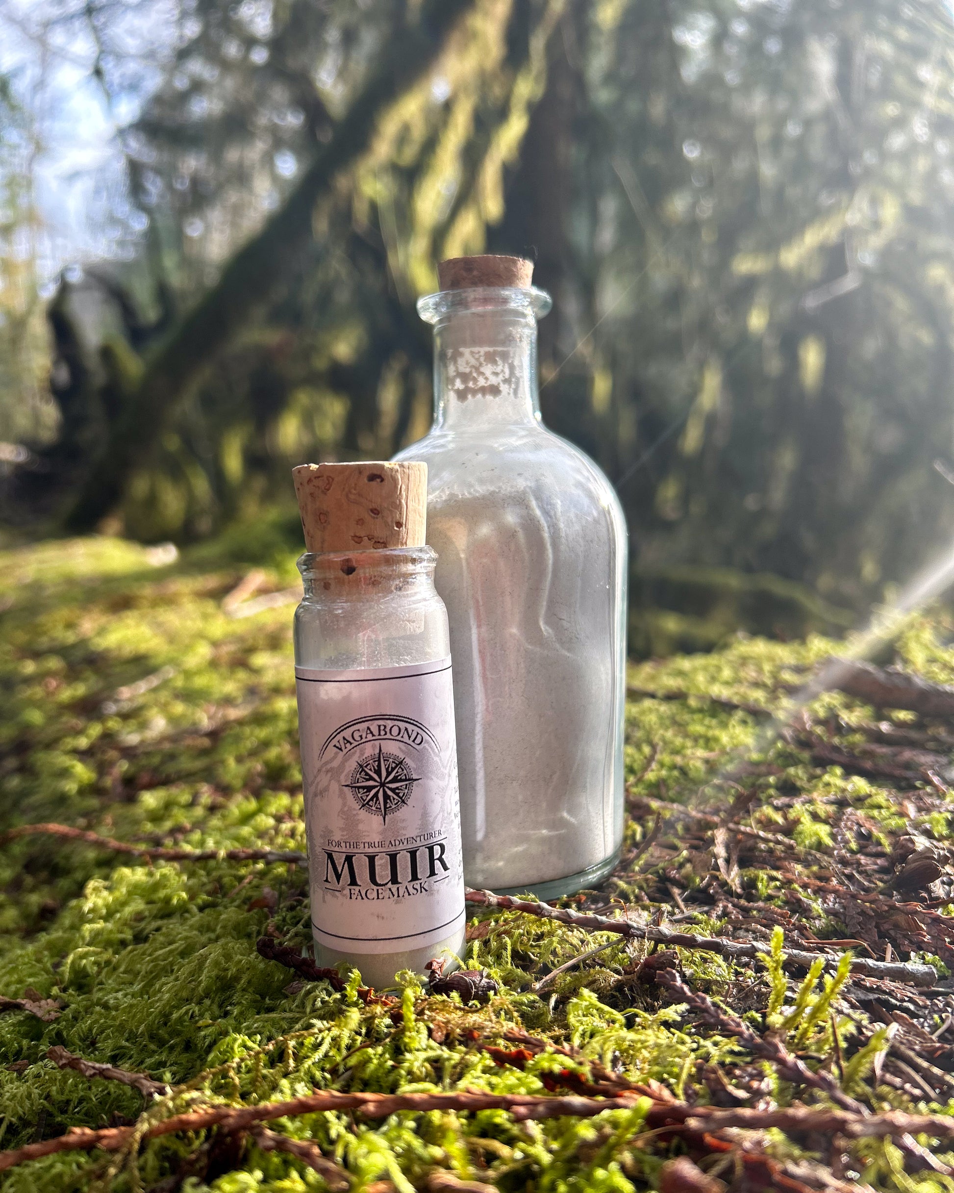 Muir natural clay mask in corked glass bottles displayed on a mossy forest floor backdrop.