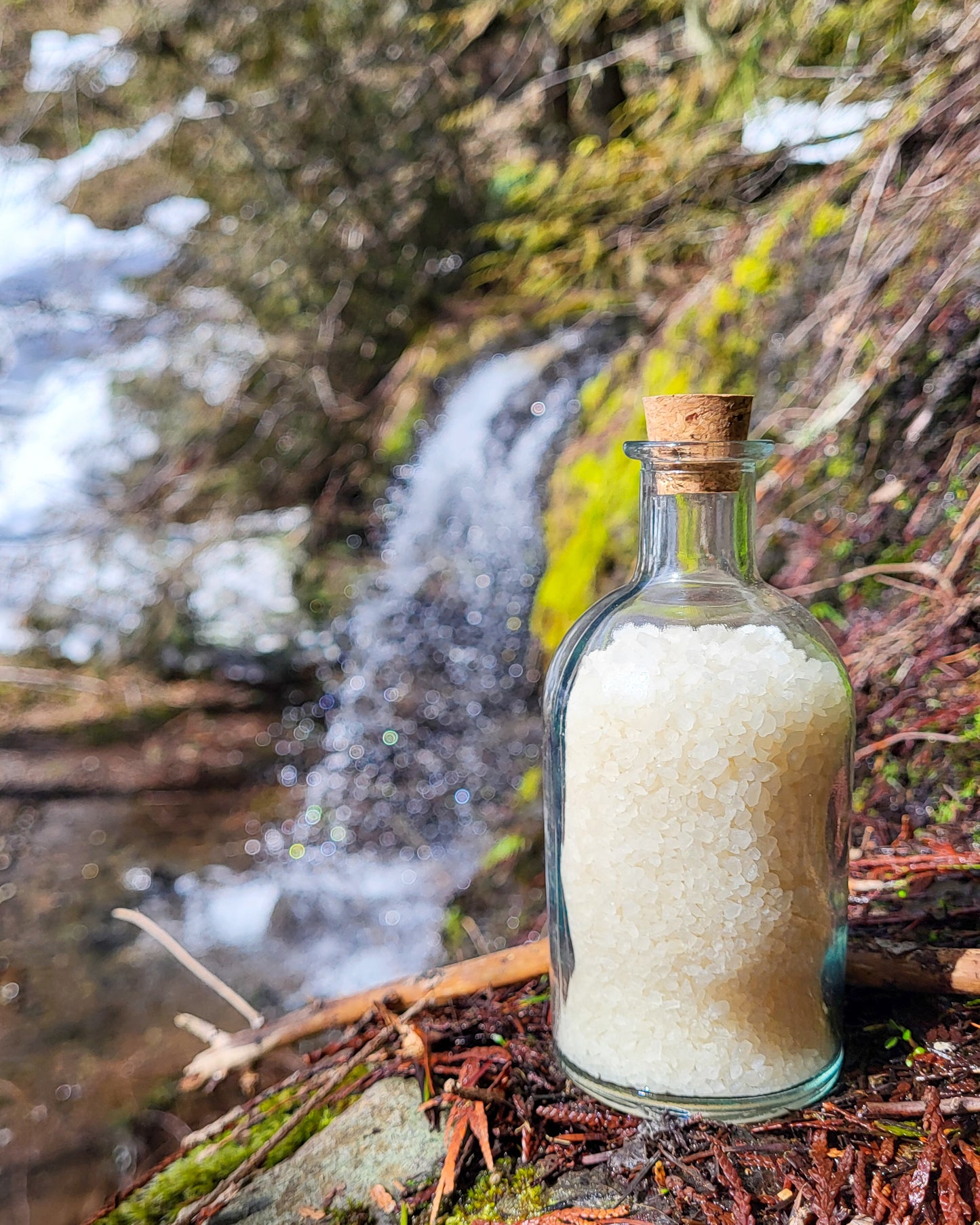 Cascadia natural bath salts lemon citrus scent in corked glass bottle displayed against a blurry waterfall hillside backdrop.